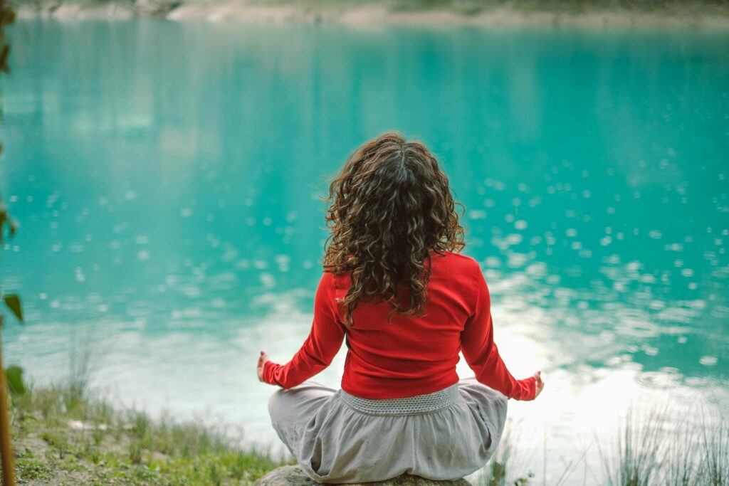 A woman sits peacefully by a calm, turquoise lake, practicing meditation as part of her wellness routine. She is dressed in a red sweater and gray pants, sitting cross-legged on a rock, embracing mindfulness and relaxation in nature. The serene scene highlights the importance of a balanced wellness routine for mental and emotional well-being.